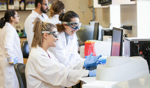 Two students wearing lab coats and safety goggles work together at laboratory equipment while other students work in the background.
