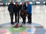 Four Ontario Agricultural College alumni from the Class of 1980 pose on a curling rink during a reunion in April 2024, holding curling brooms and an OAC Aggies jacket, with a curling stone on the ice.