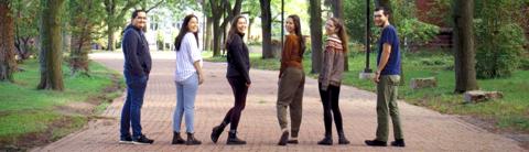A group of Indigenous students walking together on a tree-lined campus pathway, smiling and looking back towards the camera, representing community and inclusivity at the University.