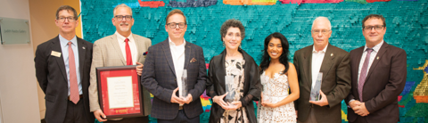 Group photo of award recipients and University of Guelph Alumni Association at the Art Gallery of Guelph. Seven people stand side by side, some holding awards and framed certificates, with a colourful mosaic background