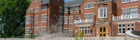Macdonald Hall at the University of Guelph, a historic red-brick building with large windows and a decorative fountain in the foreground, surrounded by greenery.