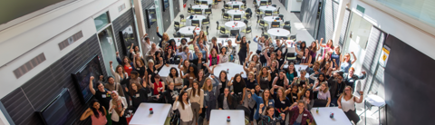 A large group of diverse women at the RISE Conference gathered in a bright, modern atrium, raising their fists in a show of empowerment and solidarity. The event setup includes round tables, creating a welcoming atmosphere for networking and discussion.