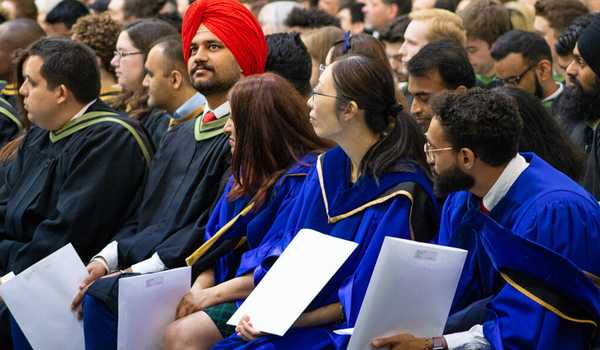 University of Guelph graduate students in blue and black robes sit during convocation, holding certificates, with a diverse group of students present.