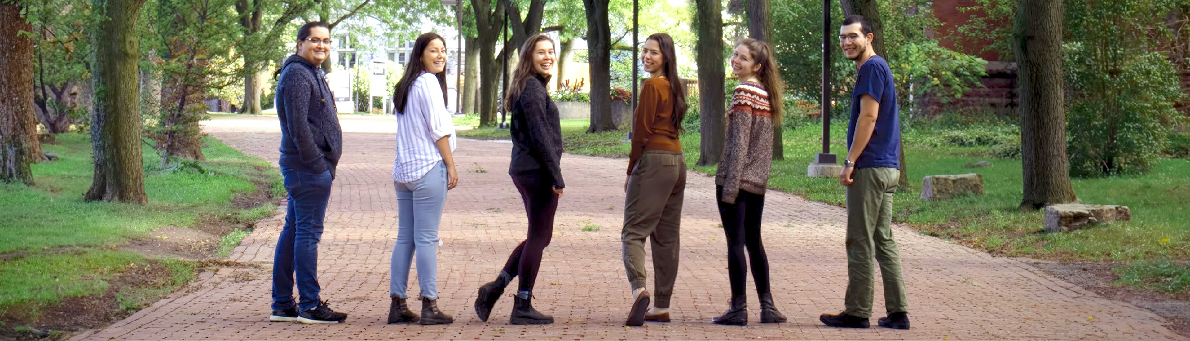 A group of Indigenous students walking together on a tree-lined campus pathway, smiling and looking back towards the camera, representing community and inclusivity at the University.