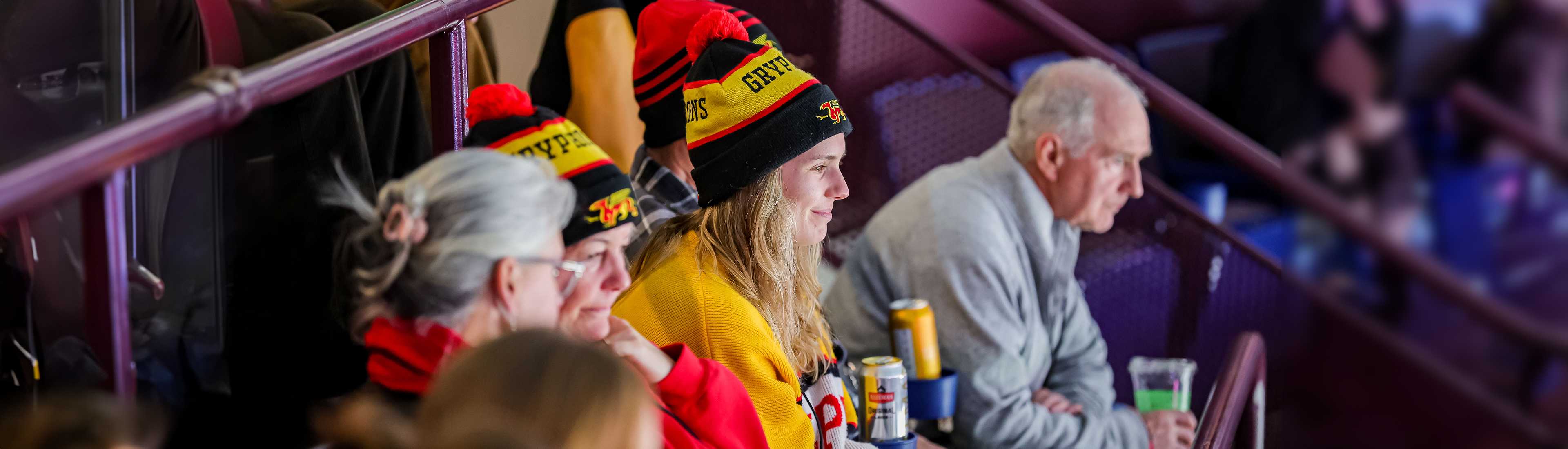 Group of enthusiastic hockey fans wearing Gryphons hats and yellow sweaters, watching a game intently from the stands. The central figure is a young woman smiling, with a soda can in front of her.