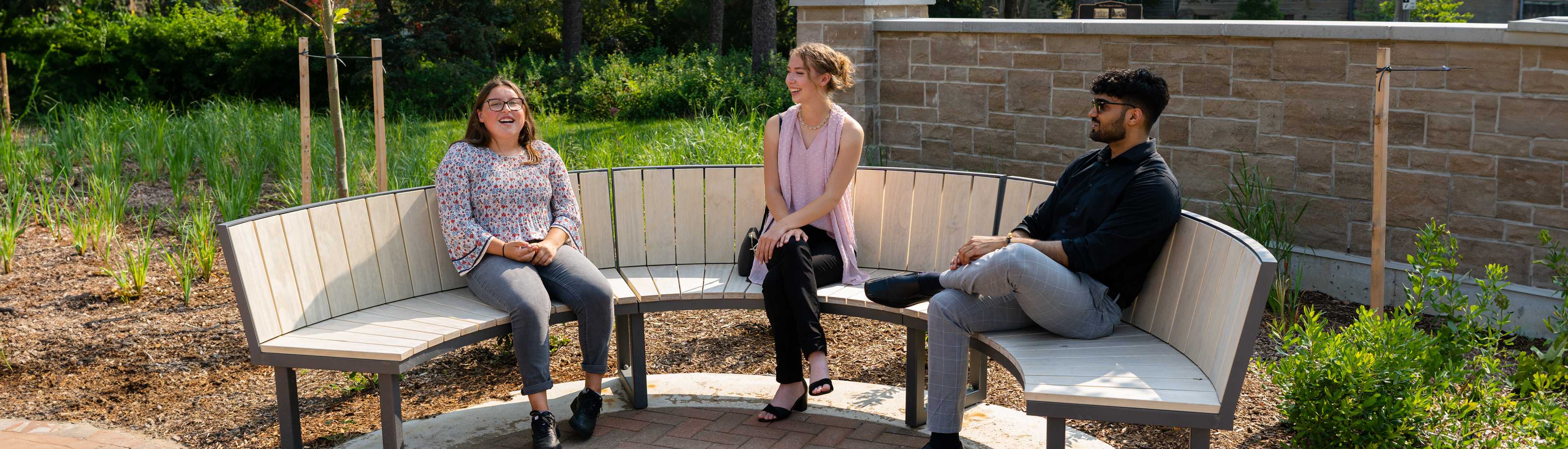 Three students sitting and chatting on a curved wooden bench in a garden area at the University of Guelph