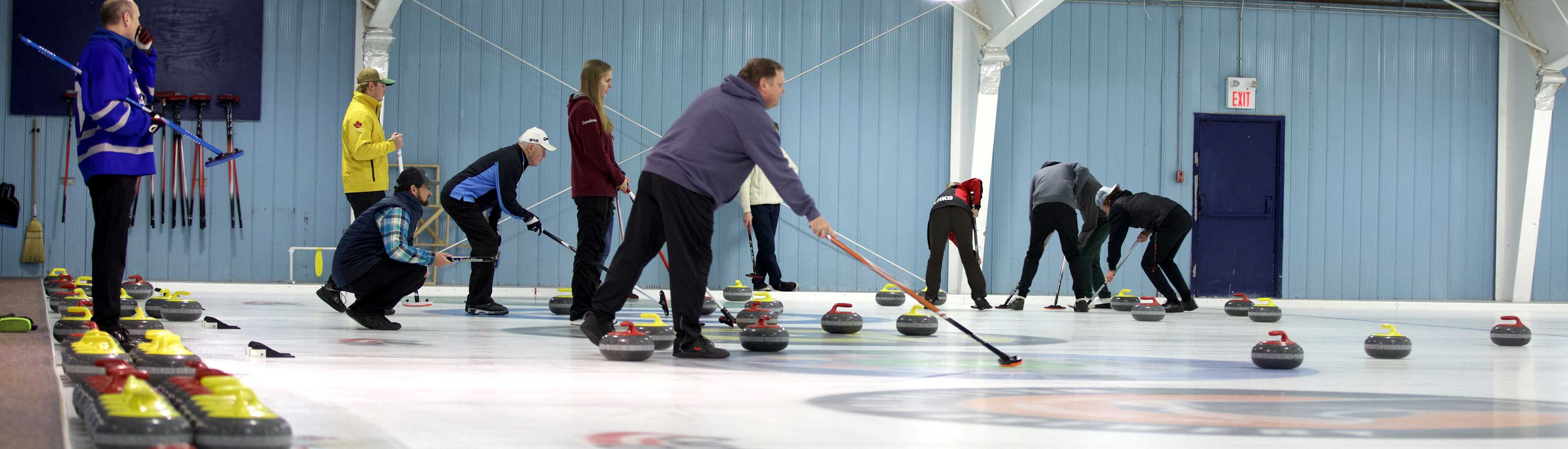 Participants at the OAC AA Bonspiel curling event, preparing and delivering stones on the ice with equipment and multiple stones arranged along the sidelines.