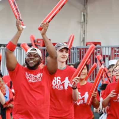 Group of university of Guelph students wearing red shirts and cheering during an outdoor orientation event, holding inflatable red sticks and smiling.