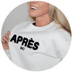 A woman wearing a white sweatshirt with bold black embroidered text reading "APRÈS SKI," smiling and holding the edge of her sweater.