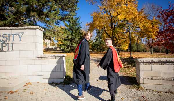 Two University of Guelph graduates in academic robes walk toward the campus on a sunny fall day, with colorful autumn trees in the background.