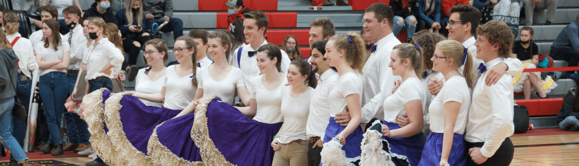 A group of young dancers in coordinated outfits pose together at College Royal at the University of Guelph. The women wear white tops and long, flowing purple skirts with lace trim, while the men wear white shirts, some with bow ties. They stand arm in arm, smiling. Behind them, a crowd of spectators and other participants, some wearing casual attire and others in themed outfits, are gathered in the bleachers and on the gym floor.