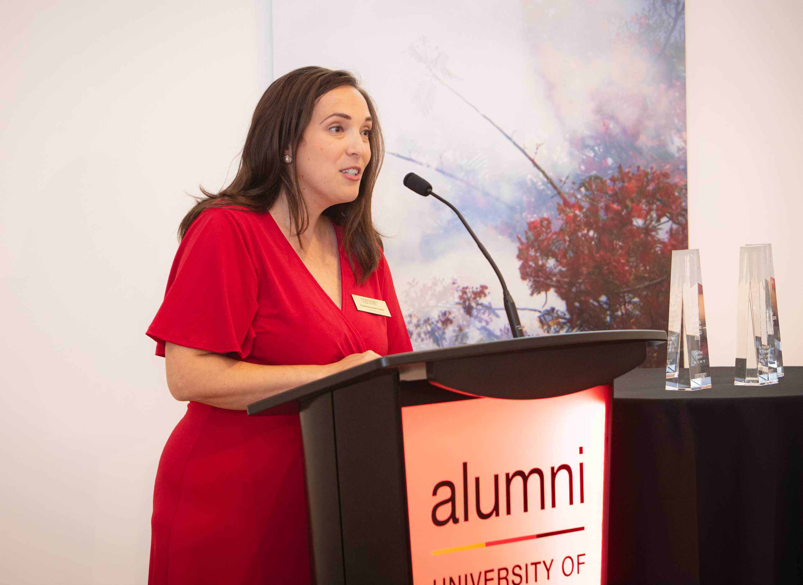 Speaker in a red dress addressing the audience at a University of Guelph alumni event, standing at a podium with an 'alumni' sign, with abstract artwork in the background.