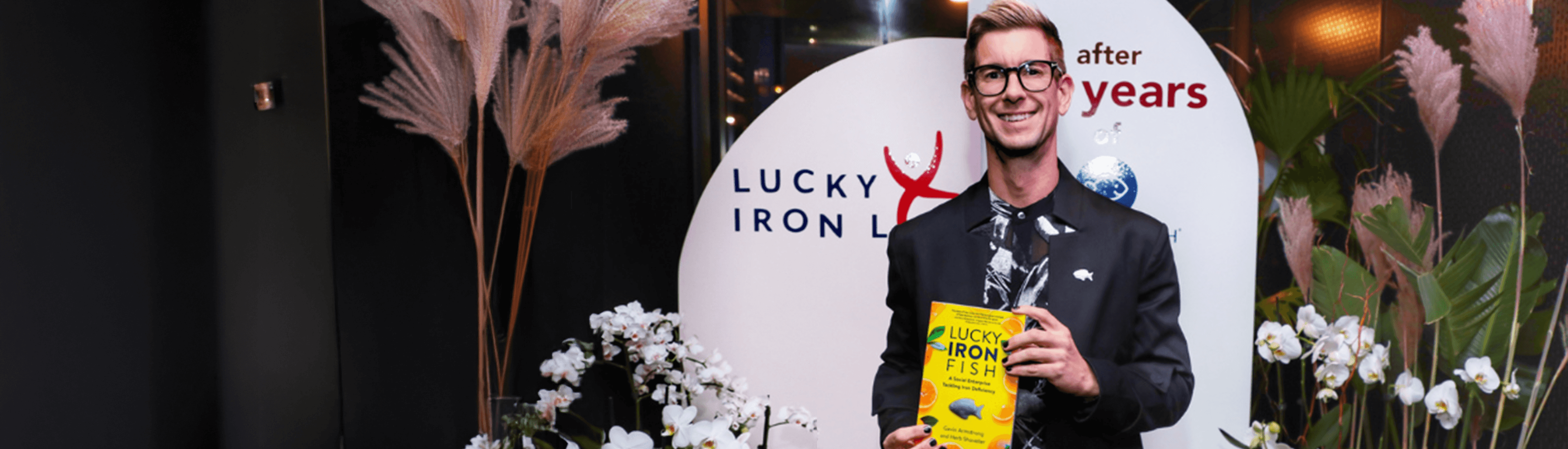 An individual in a black suit holding a Lucky Iron Fish product, standing in front of a branded backdrop with the text 'Lucky Iron Fish' and floral decorations, including pampas grass and white orchids.