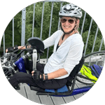 Cyndy McLean smiling while seated on a blue handcycle, wearing a white long-sleeve shirt, black gloves, sunglasses, and a bicycle helmet, with a metal railing and green trees in the background.