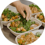 A hand adding fresh cilantro to a plant-based meal bowl filled with crispy tofu, greens, and vegetables, served with a side of dipping sauce in an eco-friendly container.