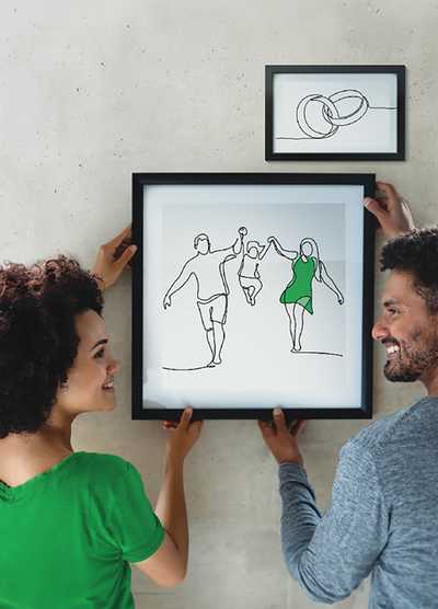 A couple hanging a framed minimalist line drawing of a family on a wall, smiling at each other, with a smaller framed drawing of wedding rings above.