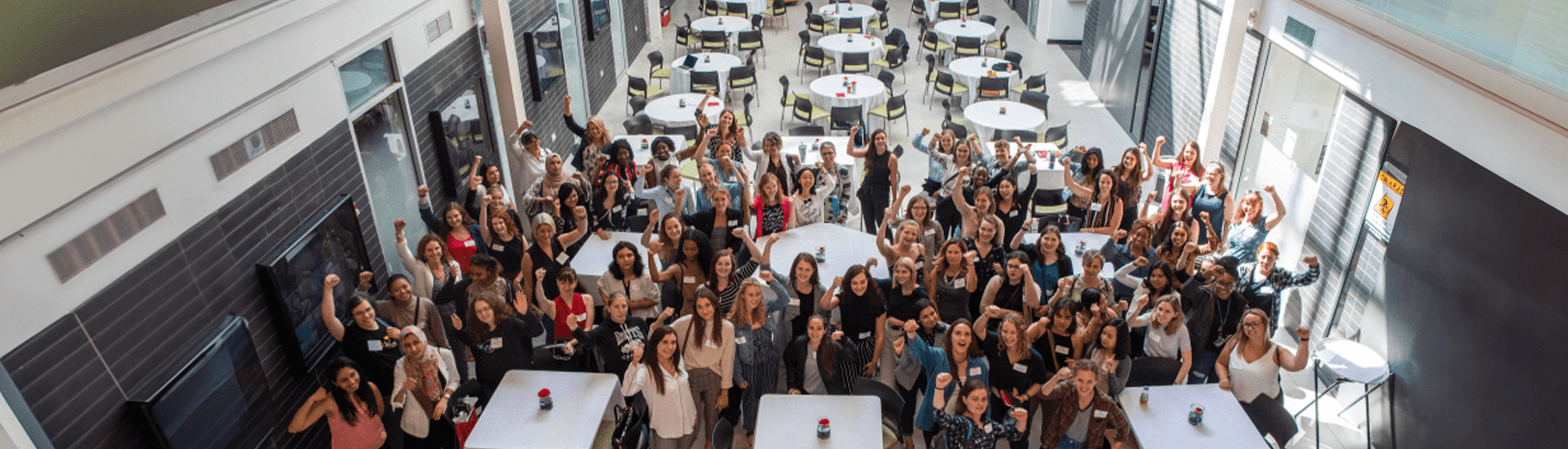 A large group of diverse women at the RISE Conference gathered in a bright, modern atrium, raising their fists in a show of empowerment and solidarity. The event setup includes round tables, creating a welcoming atmosphere for networking and discussion.